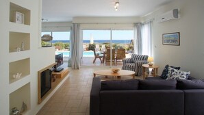 A living room with a dark sofa, a TV, and a coffee table. There is a view of a pool and outdoor furniture through large windows. - Villa for Rent in Protaras 1029 (Paralimni)