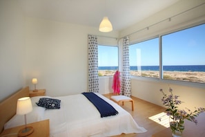 Quarto com cama, luminária de cabeceira, banqueta e vista para a praia.