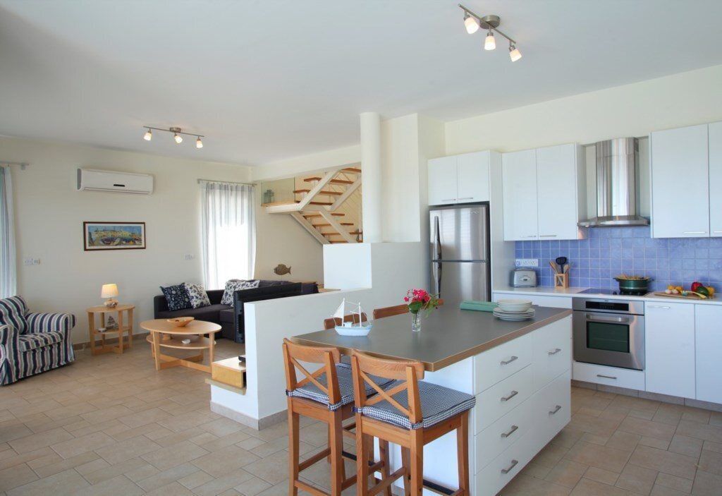 A modern kitchen with white cabinets, a stainless steel refrigerator, and a built-in oven. There is a dining area with a table and chairs, and a staircase leading to an upper level.