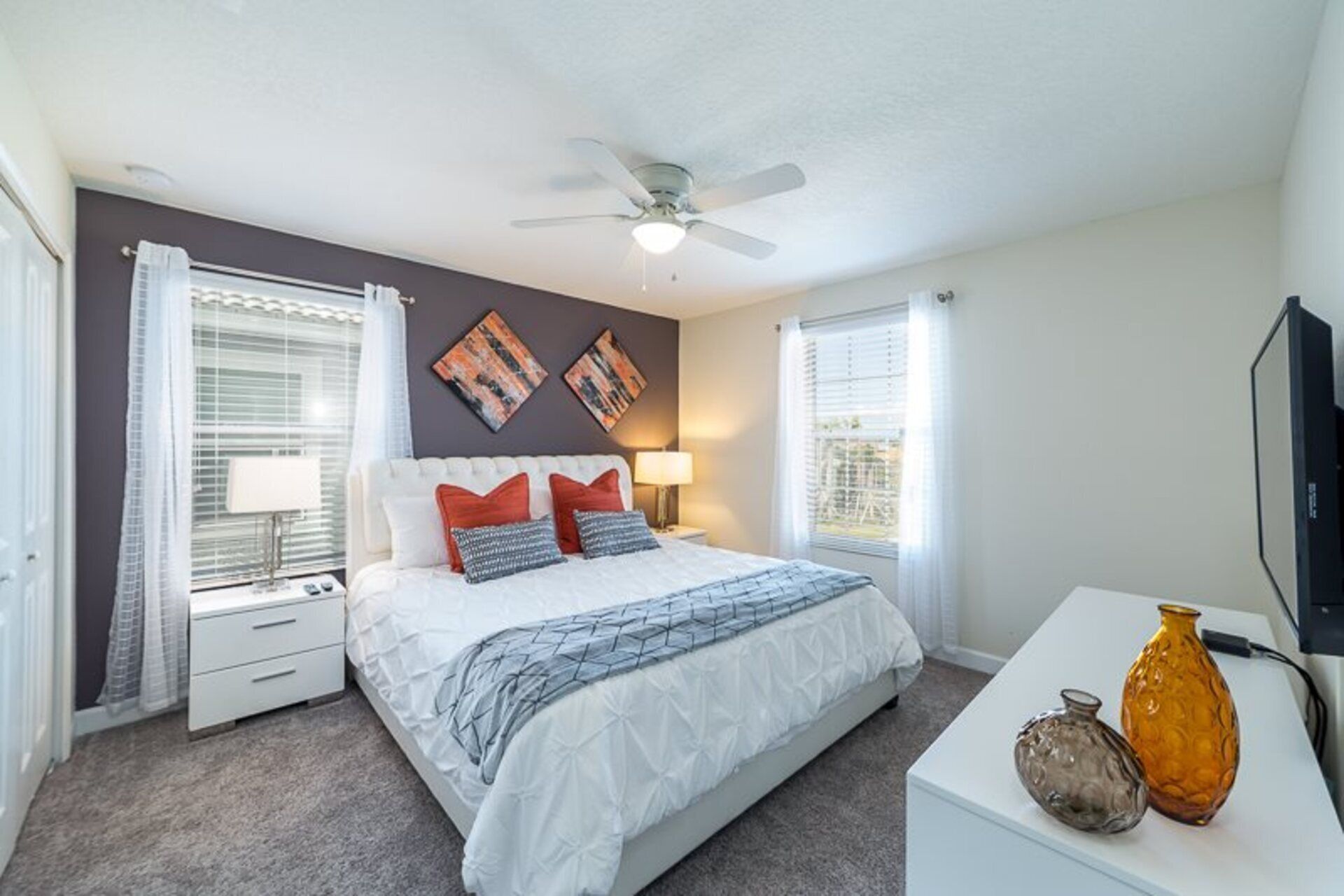 A bedroom with a bed, bedside tables, a ceiling fan, artwork on the wall, and a TV.