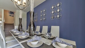 Comedor con una larga mesa dispuesta para seis, con platos, tazones y vasos blancos, y una lámpara moderna sobre la mesa.