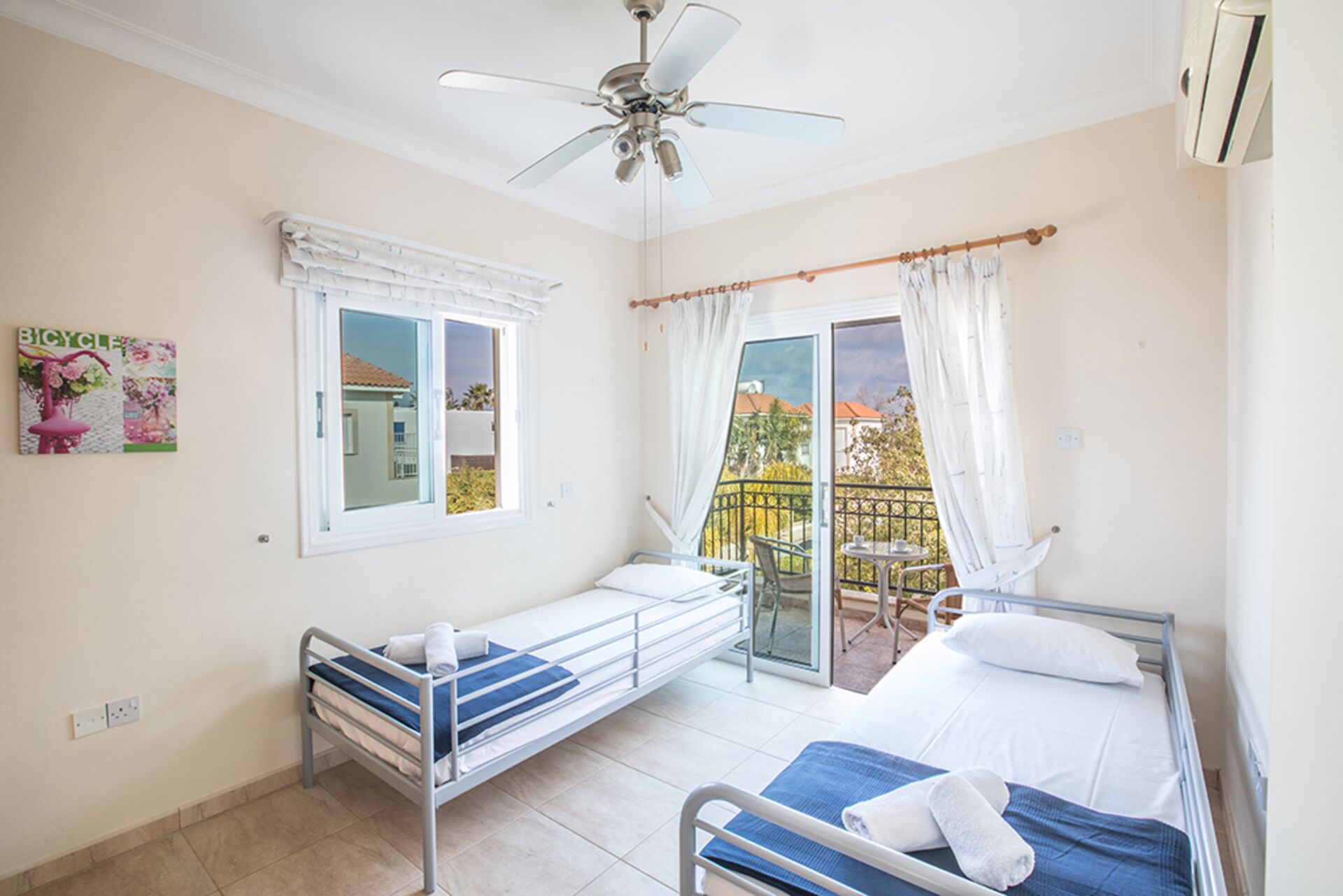 Ein Zimmer mit zwei Betten, einem Deckenventilator, einem Fenster mit Vorhängen und Blick ins Freie.
