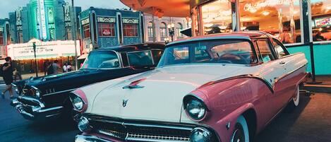 A vintage car parked in front of a diner with neon signs and a palm tree in the background.