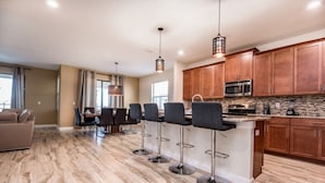 A modern kitchen with wooden cabinets, a central island with bar stools, and a dining area with a sofa and curtains.