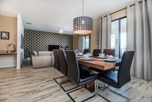 A modern dining area with a wooden table, black chairs, and a pendant light. There is a sofa and a TV in the background.