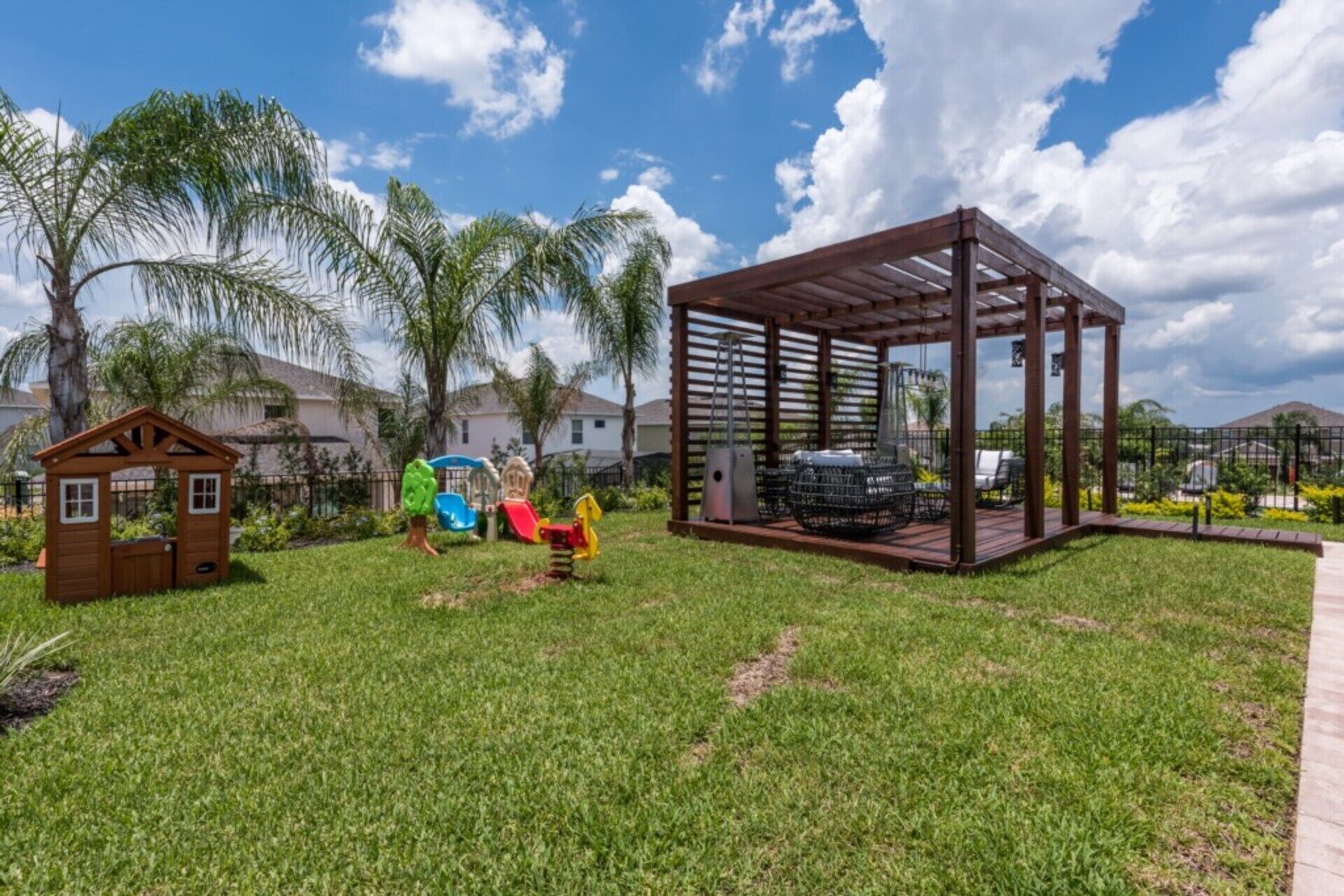 A playground with slides and a wooden playhouse, surrounded by grass and palm trees.