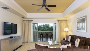 A hotel room with a flat-screen TV, a ceiling fan, a sofa, a coffee table, and a balcony with outdoor seating.