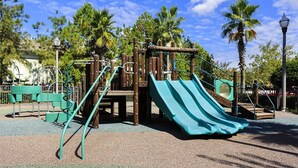 A playground with slides and climbing structures, surrounded by palm trees and a clear sky.