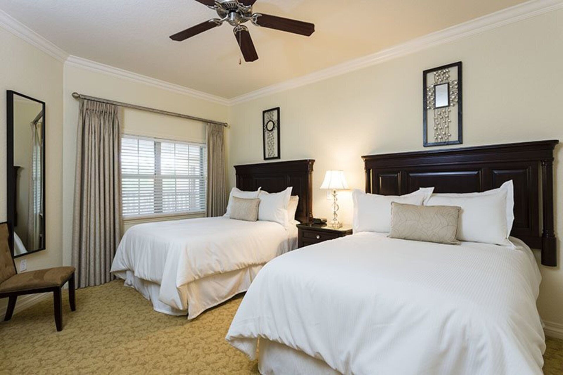 A hotel room with two beds, a ceiling fan, a window with curtains, a chair, and a nightstand with a lamp.