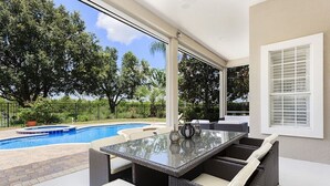 A covered outdoor patio with a glass-top table and wicker furniture, overlooking a swimming pool and lush greenery. - Rent a Luxury Villa on Reunion Resort, Minutes From Disney, Orlando Villa 3539 (Kissimmee)