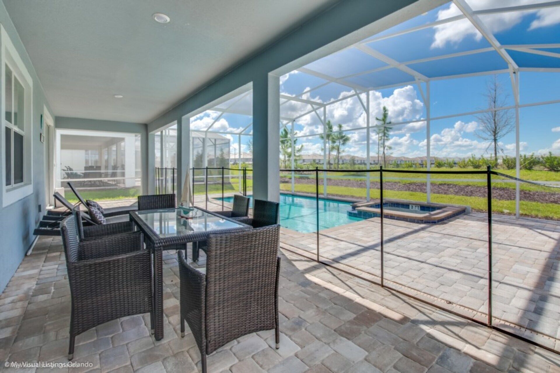 Alpendre coberto com mesa de vidro e cadeiras de vime, com vista para uma piscina e área ajardinada.