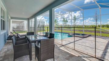 A covered patio with a glass table and wicker chairs, overlooking a swimming pool and a landscaped area.