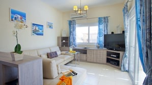 A modern living room with a sofa, a coffee table, and a TV. There is a window with curtains, a small table with a vase of flowers, and a wall-mounted air conditioner. - Cyprus Villa Near the Beach, Sotira Villa 1001 (Sotira)