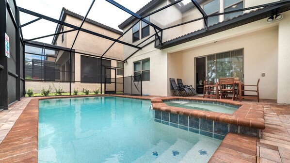 Ein Schwimmbecken mit einer gefliesten Kante, umgeben von einer Terrasse mit Holzdeck und Gartenmöbeln.