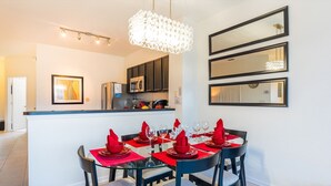 A modern kitchen with dark cabinets, a stainless steel refrigerator, and a microwave. There is a dining area with a glass table set with red napkins and wine glasses.