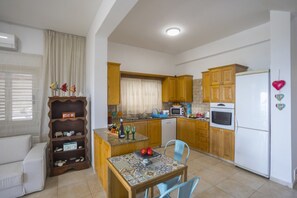 A compact kitchen with wooden cabinets, a white refrigerator, a microwave, and a dining area with a table and chairs. - Villa for Rent in Larnaca 101 (Meneou)