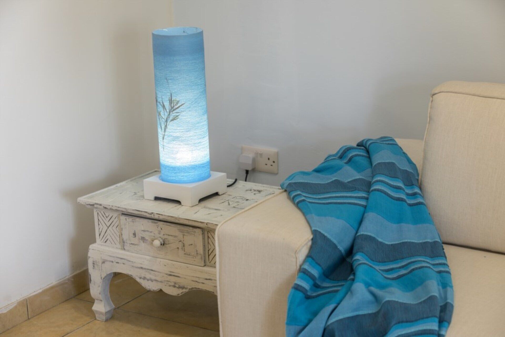 A white nightstand with a lamp, a power outlet, and a blue blanket on a beige sofa.