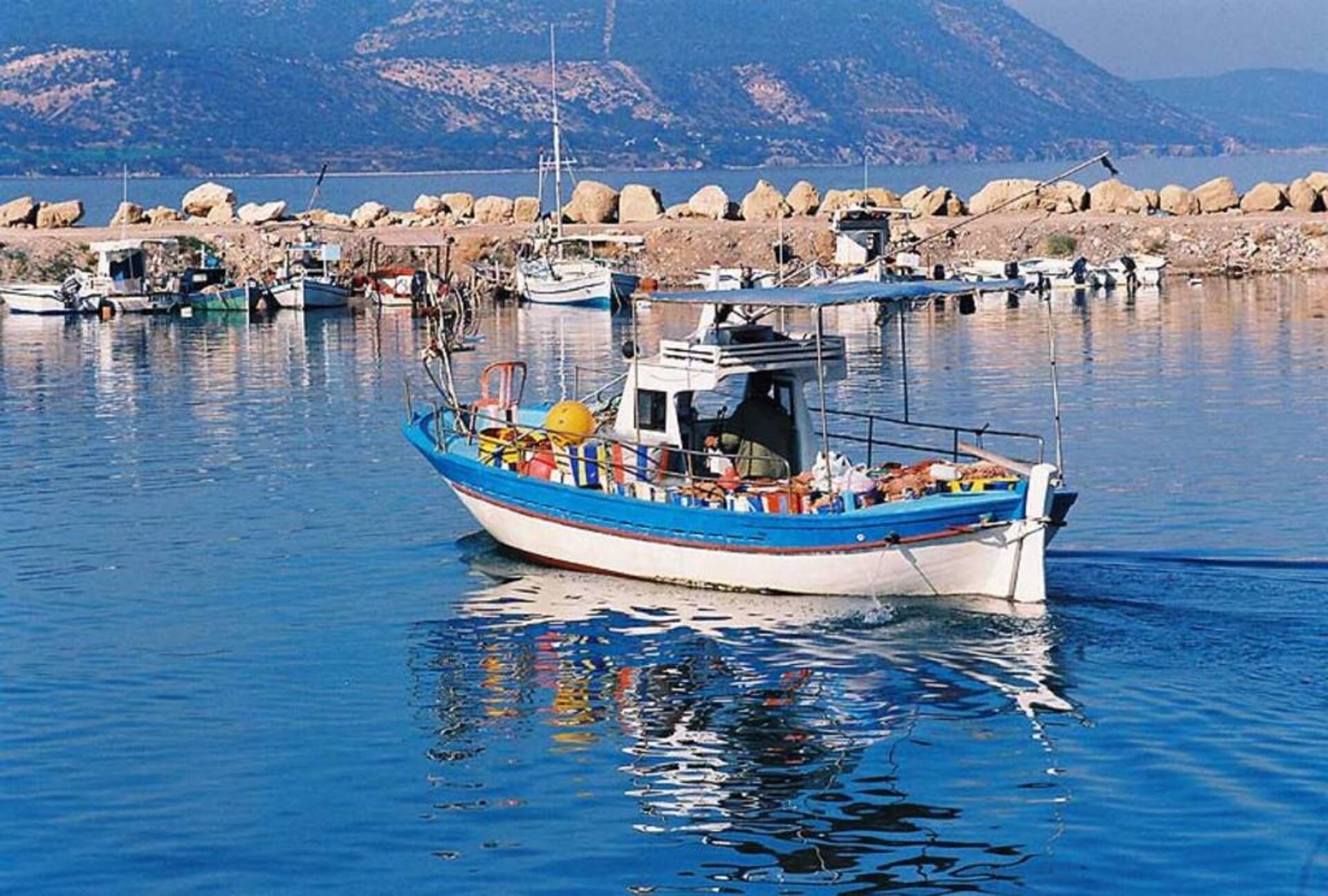 Un petit bateau doté d’une cabine et de divers équipements, naviguant sur l’eau, entouré d’autres bateaux et avec un paysage rocheux en arrière-plan.