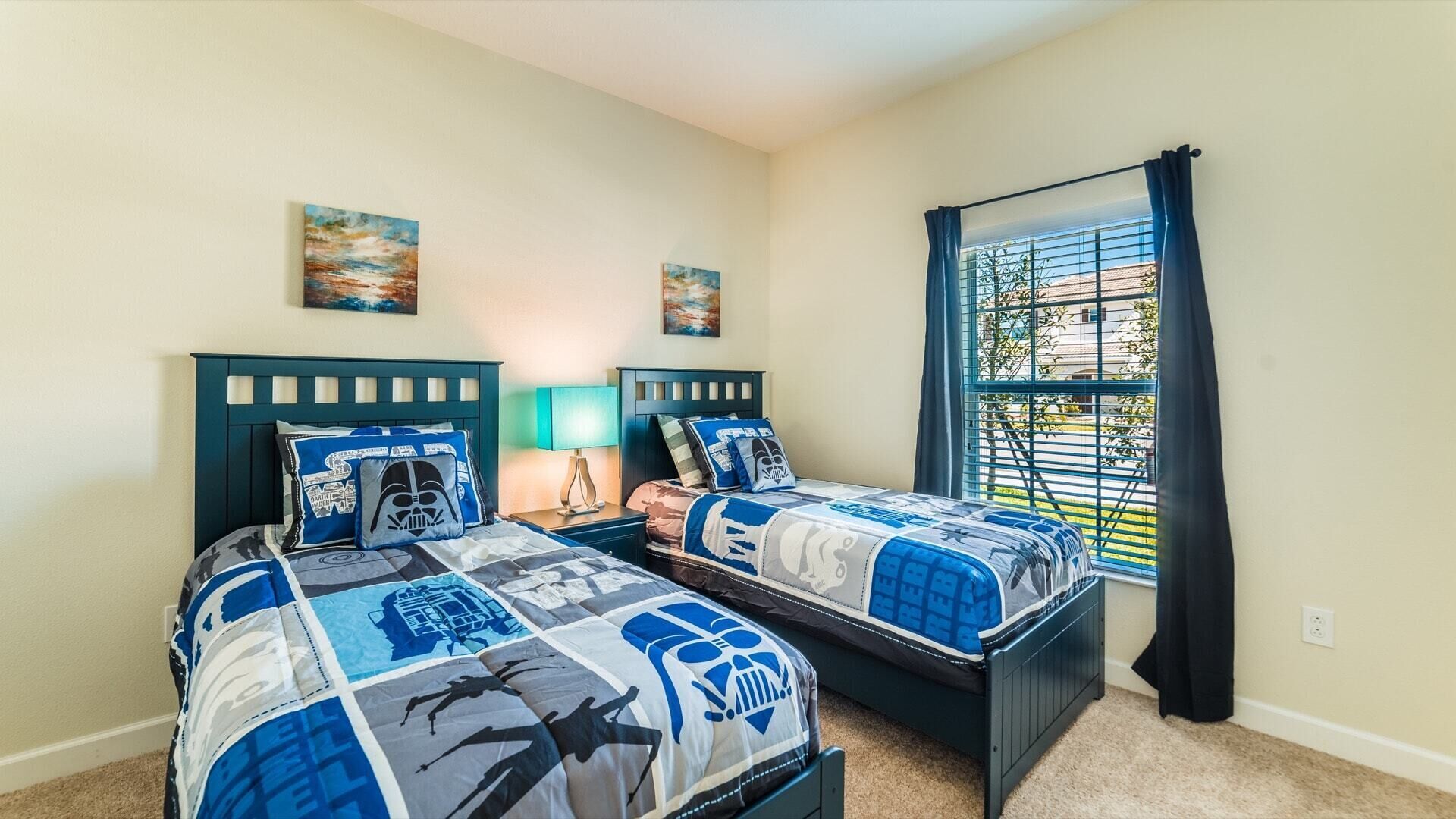 A bedroom with two beds, each with Star Wars themed bedding, a window with blinds, and two paintings on the wall.