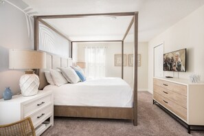 Un lit à baldaquin avec un cadre en bois, une table de chevet blanche avec des tiroirs, une lampe et des coussins décoratifs. Une télévision est posée sur un meuble en bois, à droite.