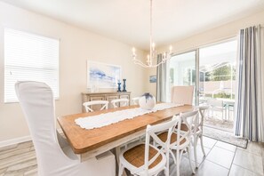 Un coin repas avec une table en bois, des chaises blanches, un lustre et une vue à travers une porte vitrée.