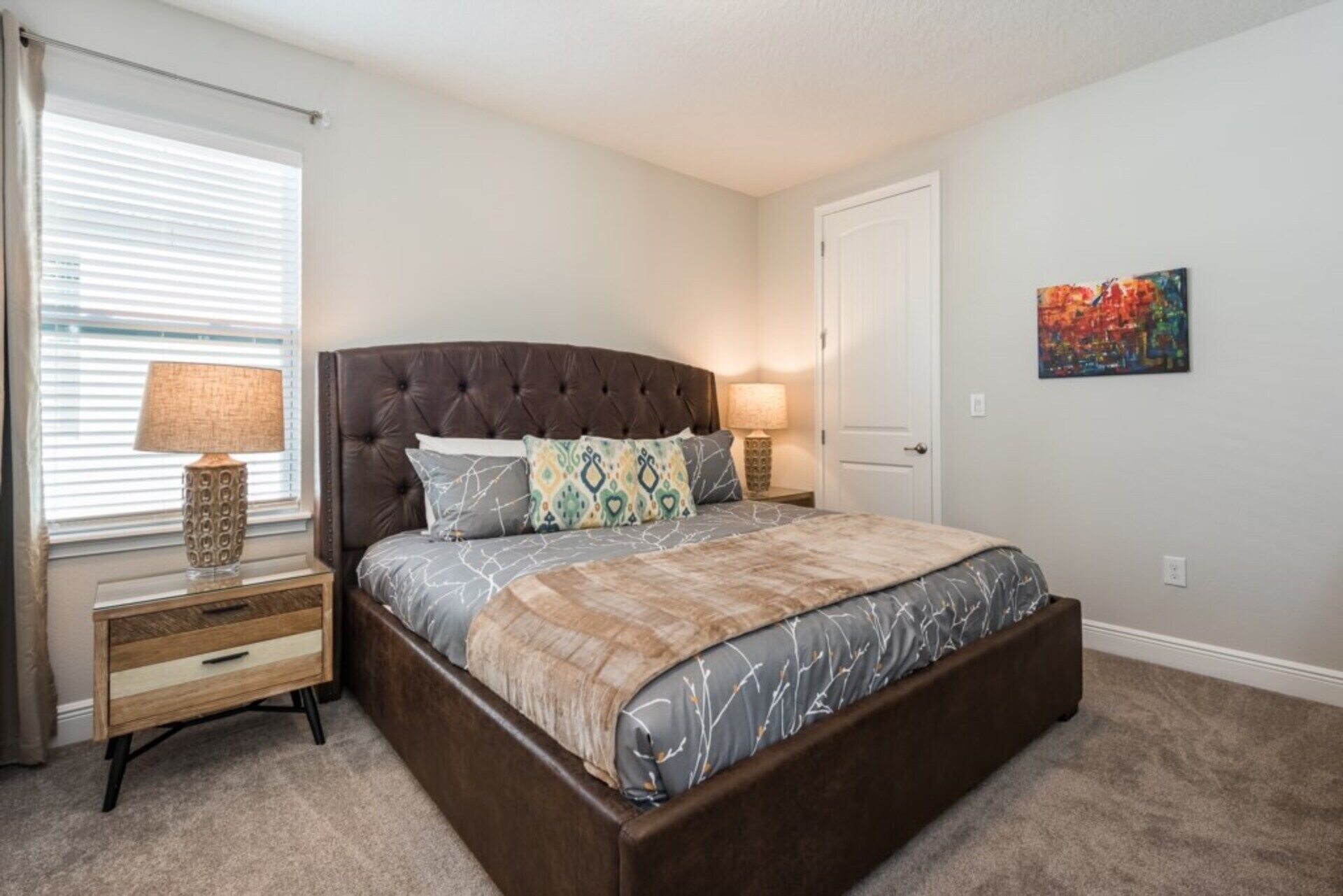 A bedroom with a large bed, a nightstand, a lamp, a door, and a painting on the wall.