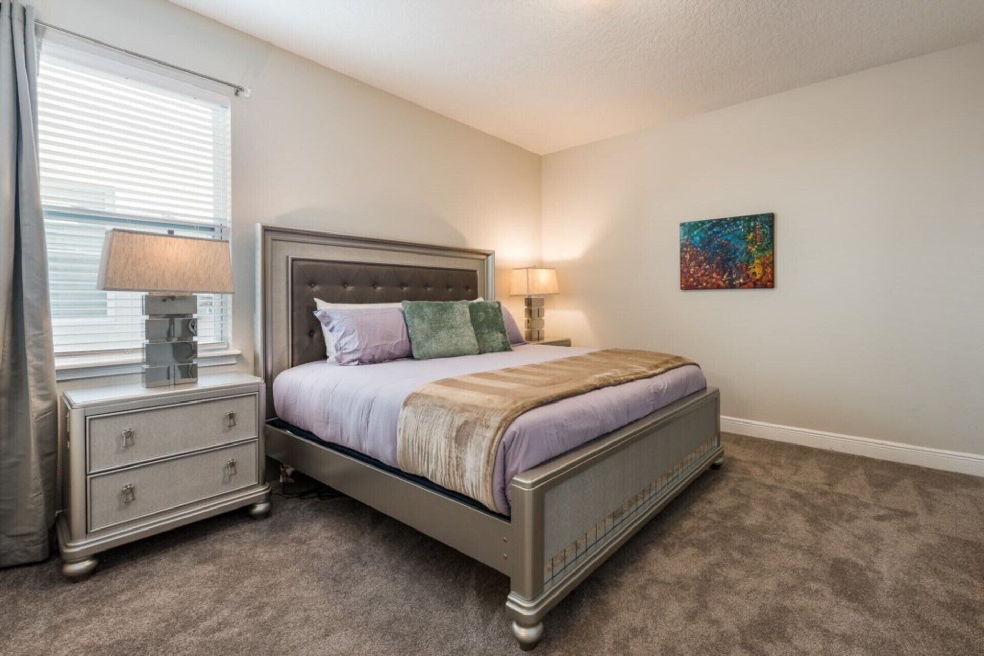 A bedroom with a bed, two bedside tables with lamps, a dresser, a window with blinds, and a colorful painting on the wall.