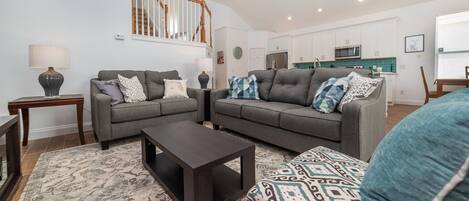 Una sala moderna con dos sofás grises, una mesa de centro oscura y cojines estampados. Una escalera con barandillas de madera conduce a un nivel superior.