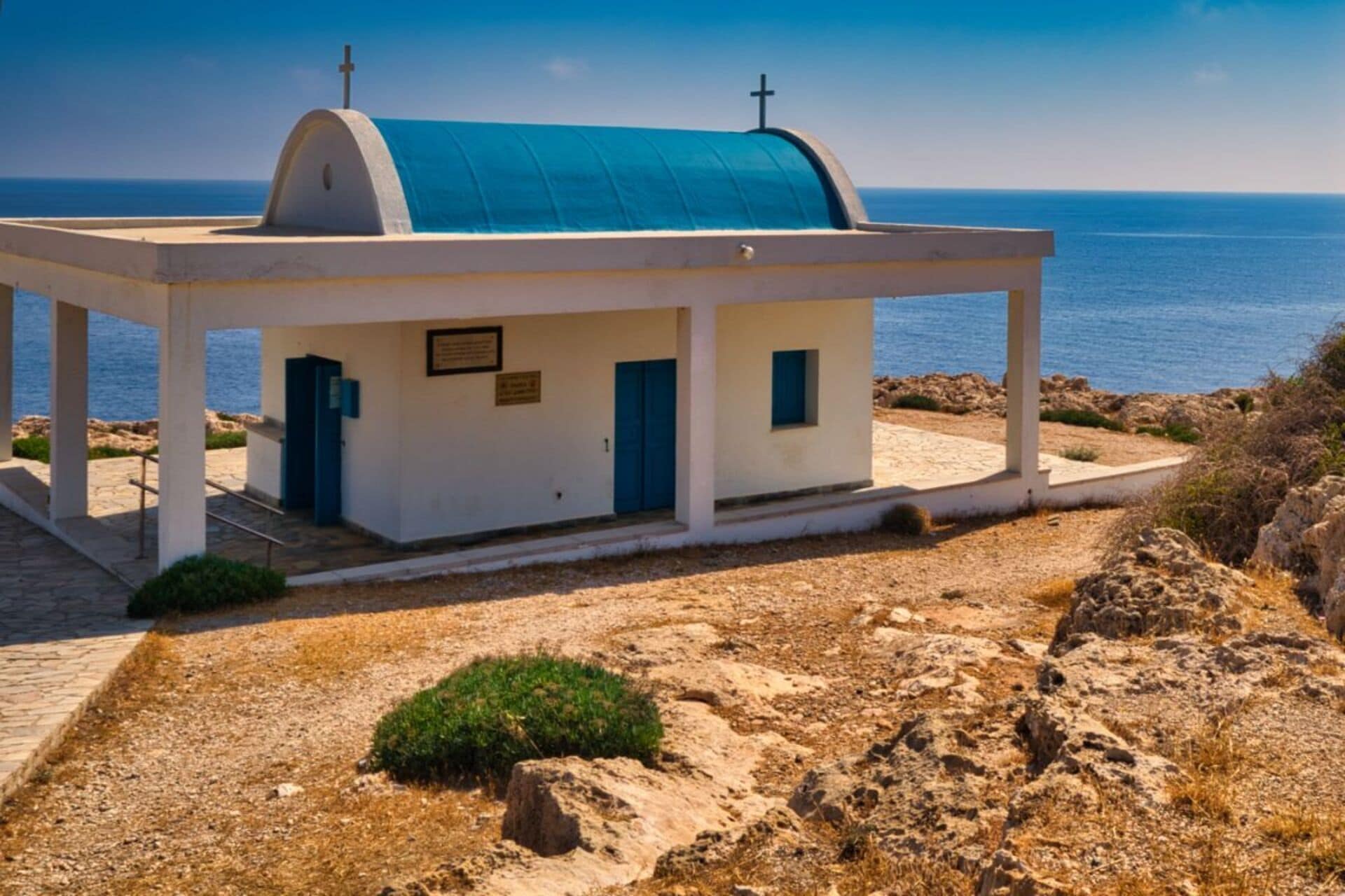 Une petite église blanche, avec un toit bleu et une croix au sommet, située sur un terrain rocheux et offrant une vue sur la mer.