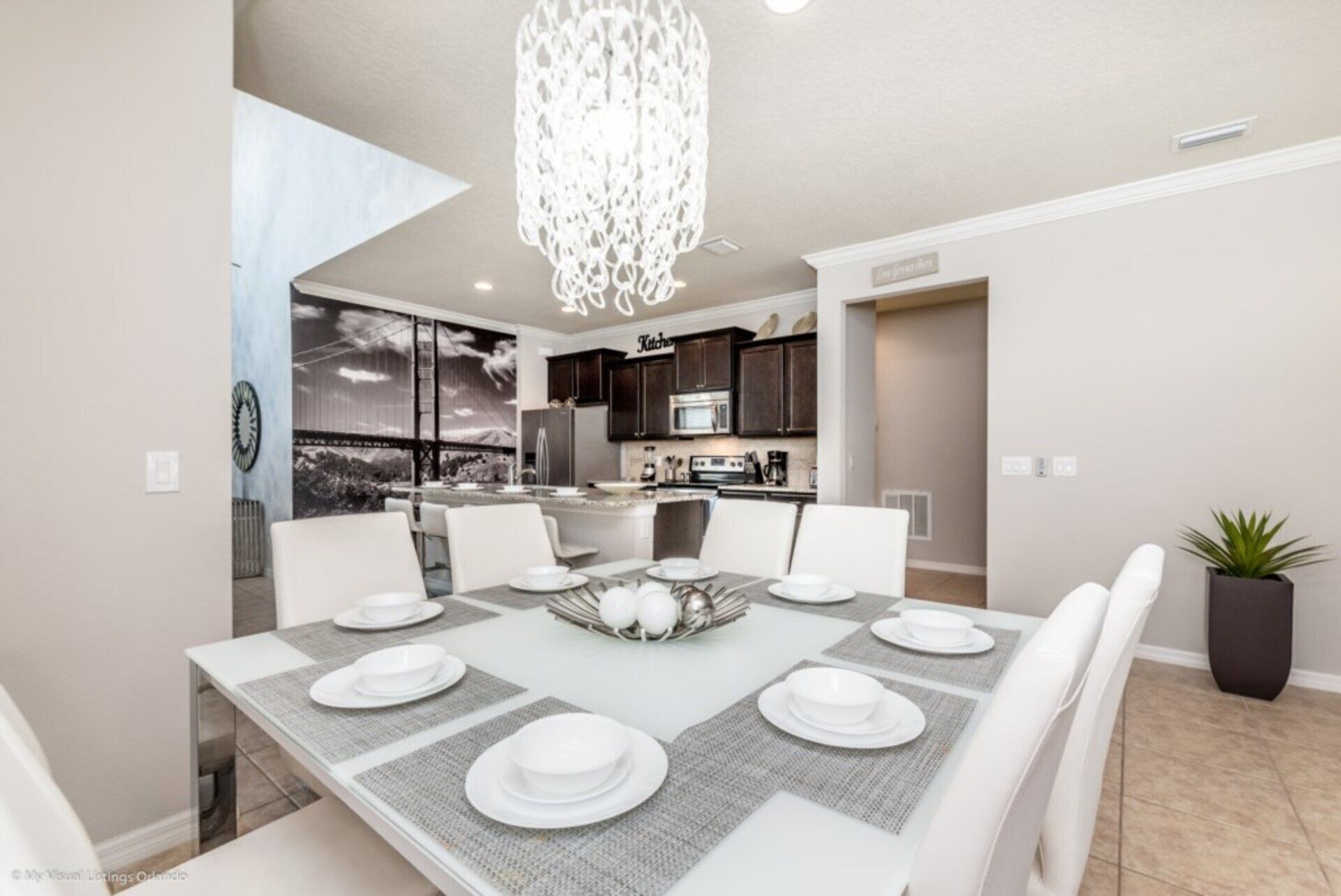 A dining area with a large table set for six, a modern kitchen in the background, and a view of a mountainous landscape through large windows.