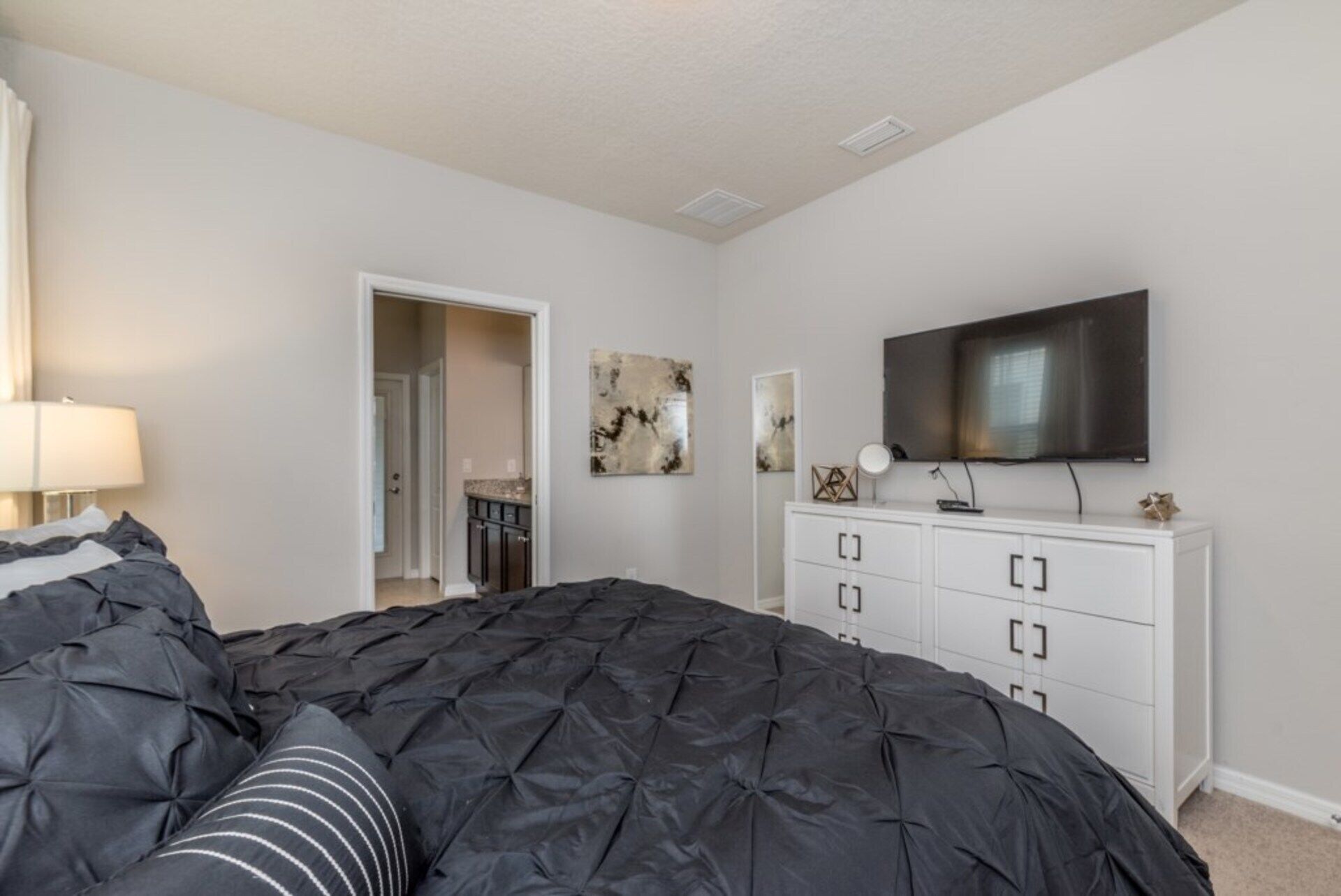 A bedroom with a large bed, a TV mounted on the wall, a dresser, and a door leading to another room.