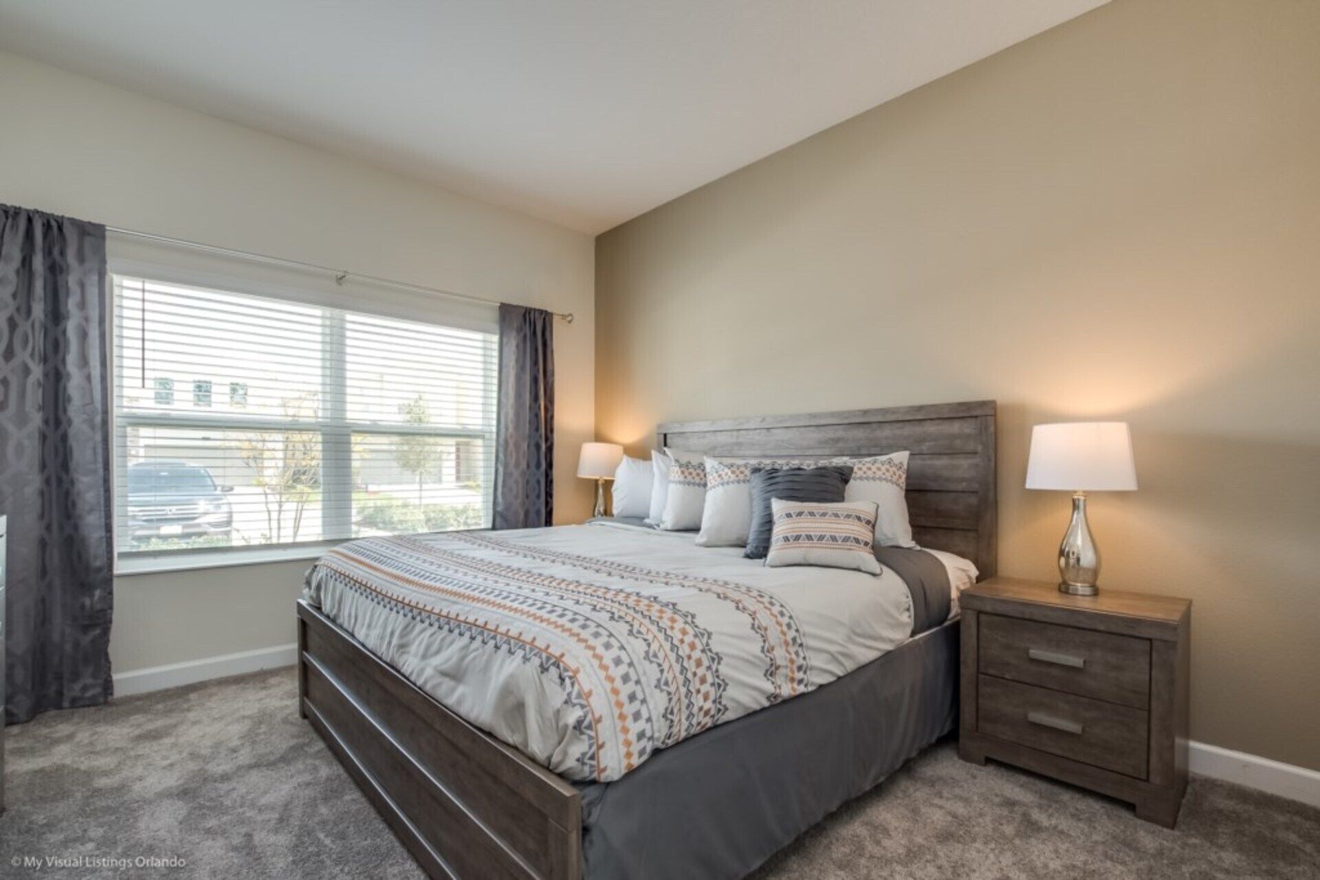 A bedroom with a large bed, two bedside tables, a window with blinds, and a view of the outside.