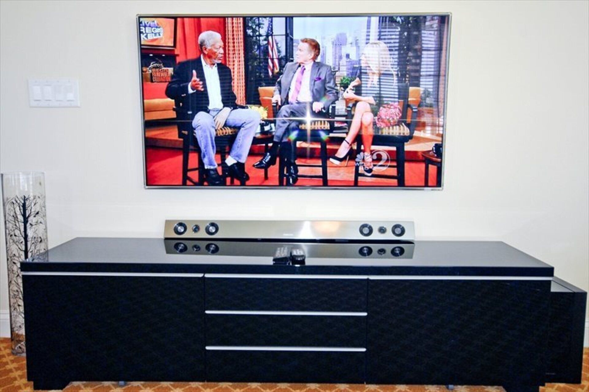 Televisor de pantalla plana montado en la pared sobre un mueble multimedia negro con varios controles.