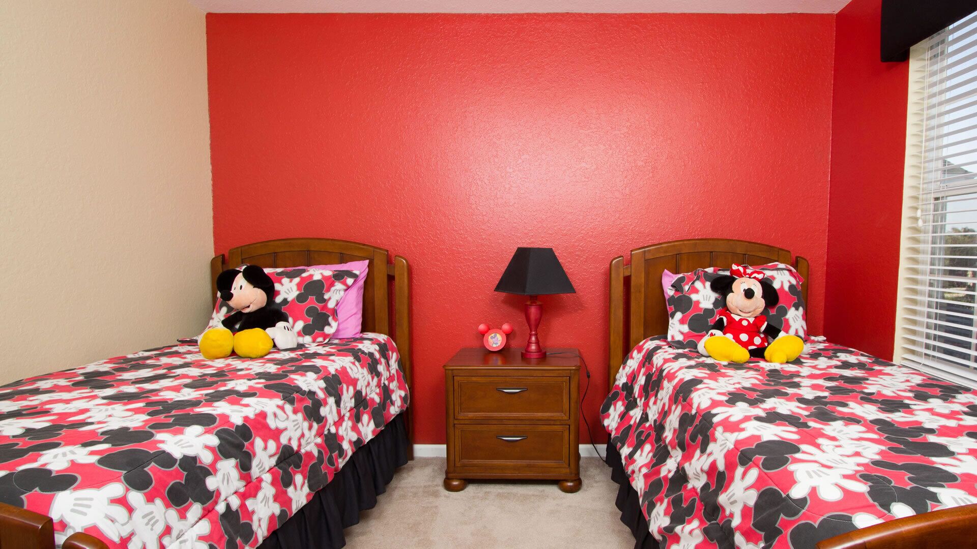 A bedroom with two beds, each with a Mickey Mouse themed bedspread, a nightstand with a lamp, and a window with blinds.