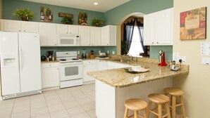 A kitchen with white cabinets, a granite countertop, a built-in oven, a microwave, and a coffee maker. There are three wooden stools at the island. - Villa in Windsor Hills Resort 1407 (Kissimmee)