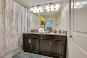 A bathroom with dark wood cabinets, a granite countertop, a large mirror with lights, and a shower with a leaf-patterned curtain.