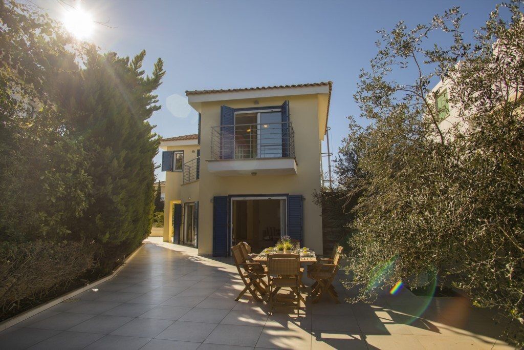 Un edificio de dos plantas con un balcón, un patio con baldosas y un área de comedor con mobiliario exterior.