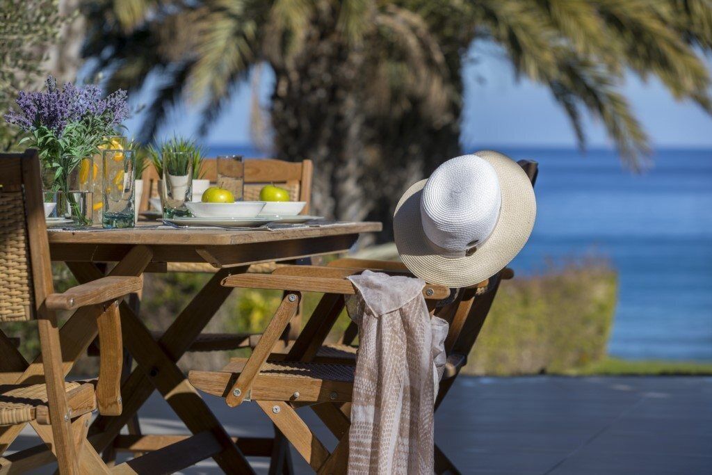 A wooden table set outdoors with a hat on the chair, a plate with lemons, and a vase with flowers.