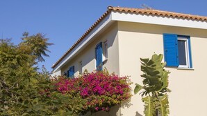 Un bâtiment aux murs blancs et aux volets bleus, entouré d’une végétation luxuriante et de fleurs roses éclatantes.