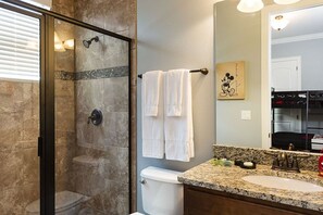 A bathroom with a glass-enclosed shower, a white toilet, a granite countertop sink, and a mirror reflecting a kitchen area. - Rent a Luxury Villa on Reunion Resort, Minutes From Disney, Orlando Villa 3556 (Kissimmee)