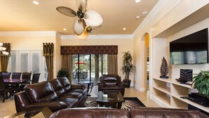 A spacious living room with a brown leather sofa, a coffee table, and a ceiling fan. There is a dining area with a table and chairs, and a TV mounted on the wall.