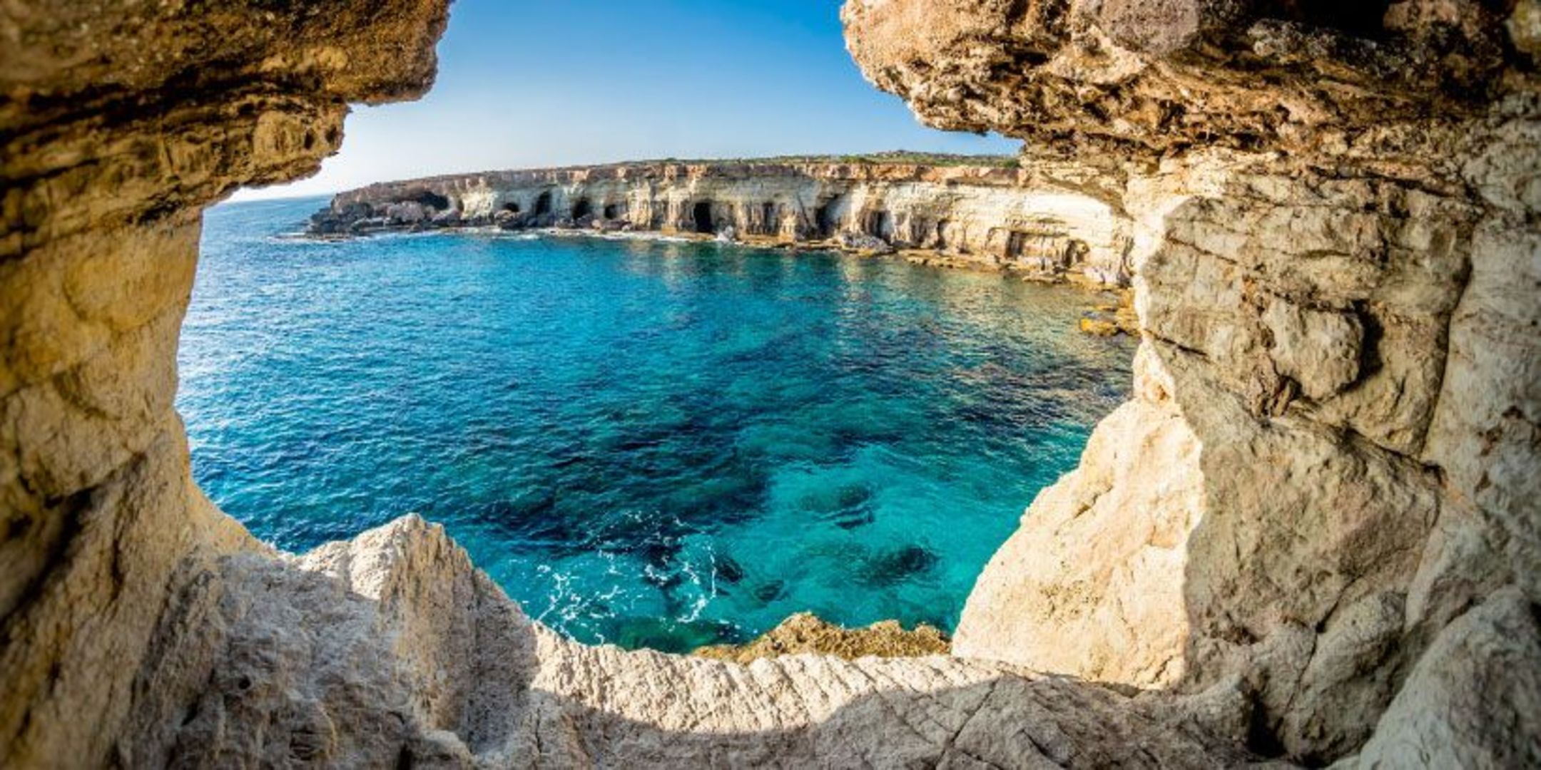 A coastal view through a natural rock arch with clear blue sea and rocky cliffs.