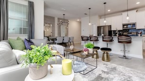 A modern living room with a sofa, coffee table, and decorative items. A dining area with chairs and a table is visible in the background. The kitchen area includes a refrigerator, microwave, and bar stools. - Rent a Luxury Townhome on Storey Lake Resort, Minutes From Disney, Orlando Townhome 3224 (Kissimmee)