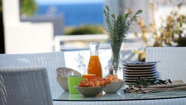 A table set with breakfast items, including croissants, a glass of orange juice, and a bottle of orange juice, with a view of the sea in the background.