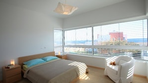 A modern bedroom with a large window, a bed, a nightstand, and a white chair.