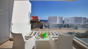 A balcony with white wicker furniture, a table with a green bowl, and a clear view of the sea and buildings in the background.