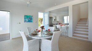 A modern dining area with a glass table surrounded by white wicker chairs, a ceiling fan, and a kitchen in the background.