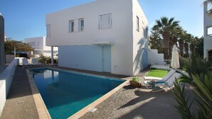A modern white building with a swimming pool, a small garden, and a clear blue sky.