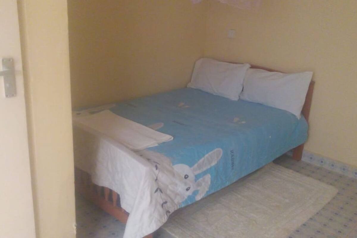 Quarto pequeno com uma cama de solteiro, cobre azul, almofadas brancas e manta branca.