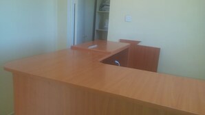 Un escritorio de madera con un estante integrado y un cajón, un enchufe eléctrico blanco en la pared, y una puerta que da a un área de almacenamiento.